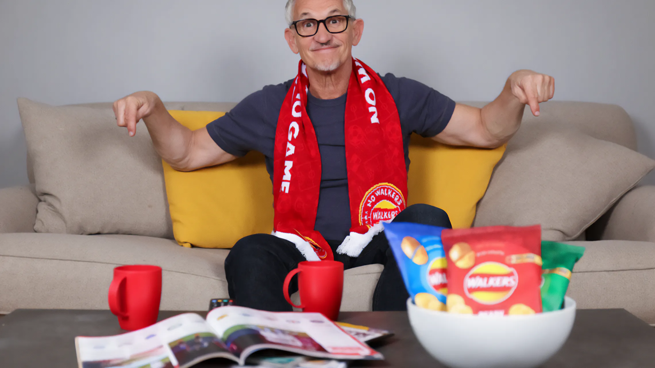 Gary Lineker sitting on a sofa with Walkers crisps, a football scarf, and red mugs during a football game.